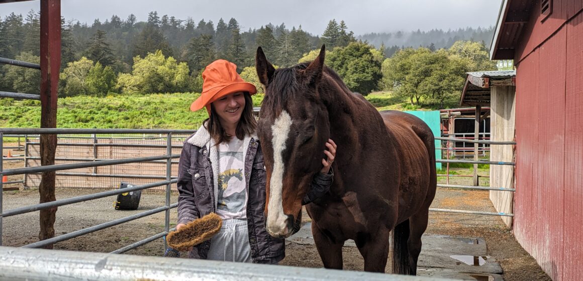 A young woman stands smiling in an orange rain hat next to a brown horse with a white stripe down his nose.