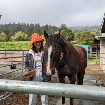 A young woman stands smiling in an orange rain hat next to a brown horse with a white stripe down his nose.