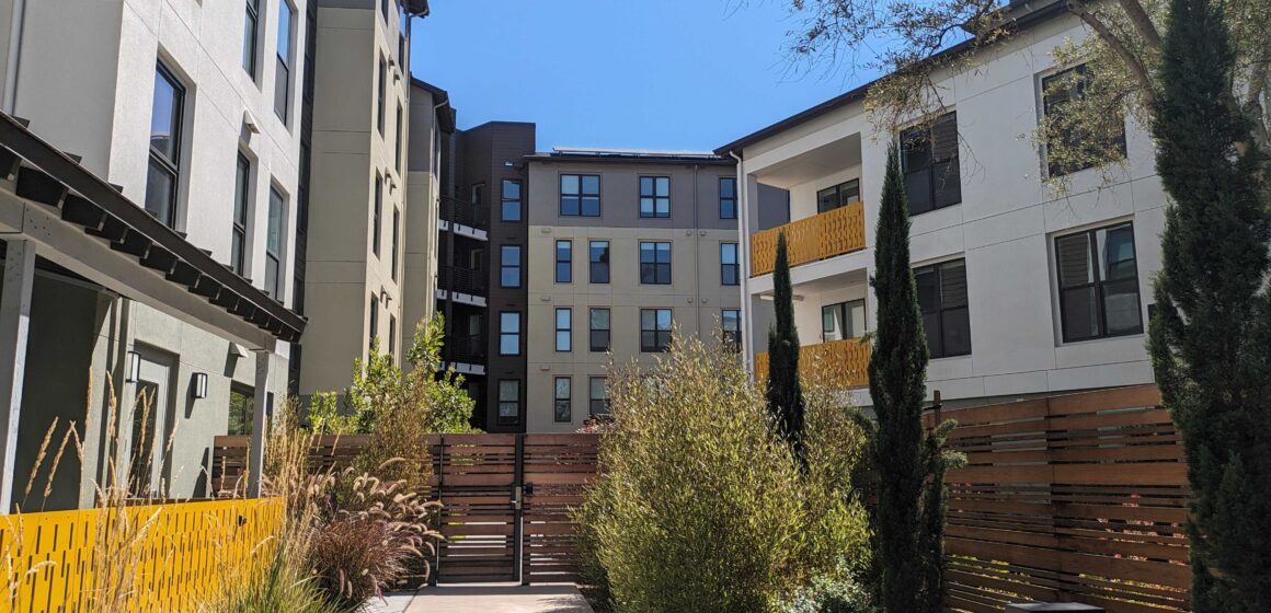 A sidewalk path leads to a gate in front of a five-story apartment building