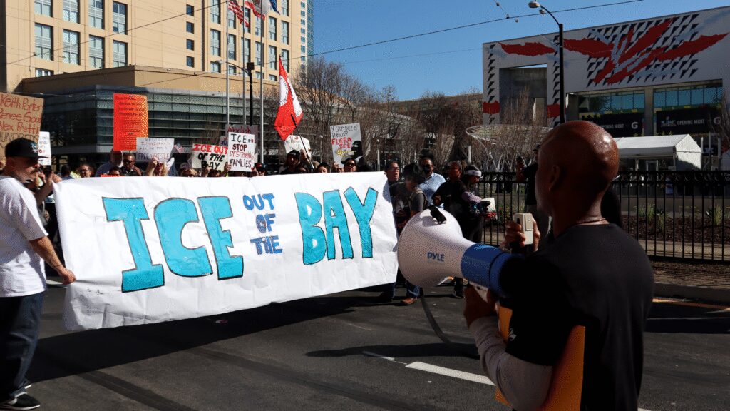 Photo of the back of a man holding a megaphone surveying a group of people standing behind a large paper banner reading "ICE OUT OF THE BAY"