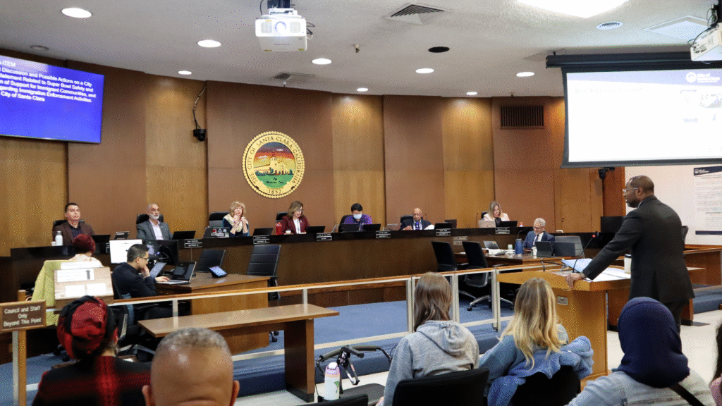 Photo of the Santa Clara City Council chambers with councilmembers sitting at their dias and City Manager Jovan Grogan, a tall Black man in a suit, giving a presentation