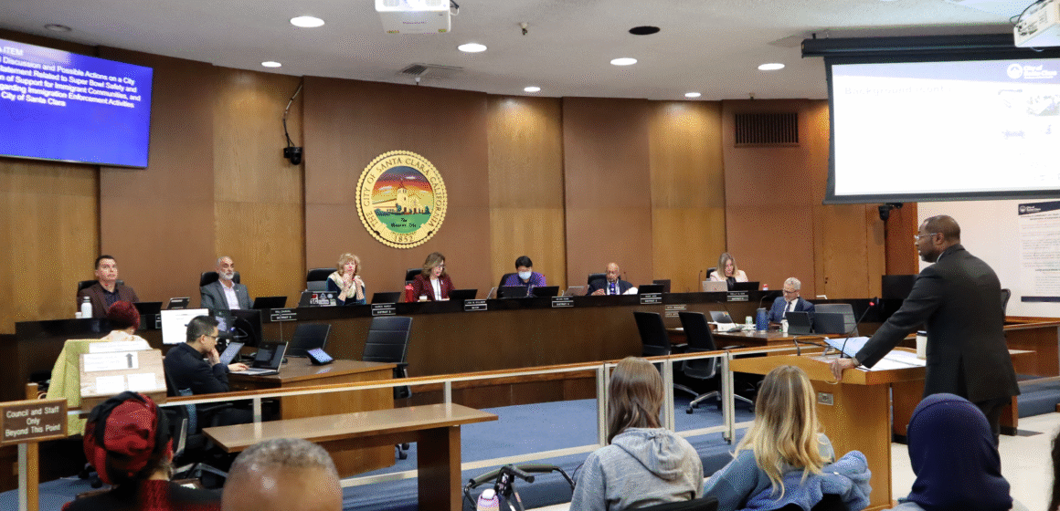 Photo of the Santa Clara City Council chambers with councilmembers sitting at their dias and City Manager Jovan Grogan, a tall Black man in a suit, giving a presentation