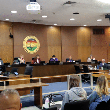 Photo of the Santa Clara City Council chambers with councilmembers sitting at their dias and City Manager Jovan Grogan, a tall Black man in a suit, giving a presentation