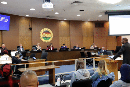 Photo of the Santa Clara City Council chambers with councilmembers sitting at their dias and City Manager Jovan Grogan, a tall Black man in a suit, giving a presentation