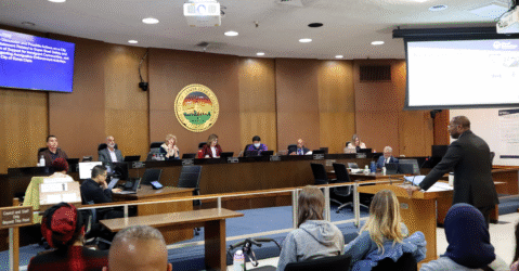 Photo of the Santa Clara City Council chambers with councilmembers sitting at their dias and City Manager Jovan Grogan, a tall Black man in a suit, giving a presentation