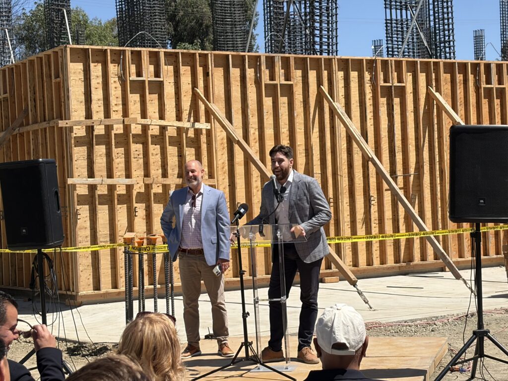 Two men speak to an audience at a construction site in Santa Clara, California